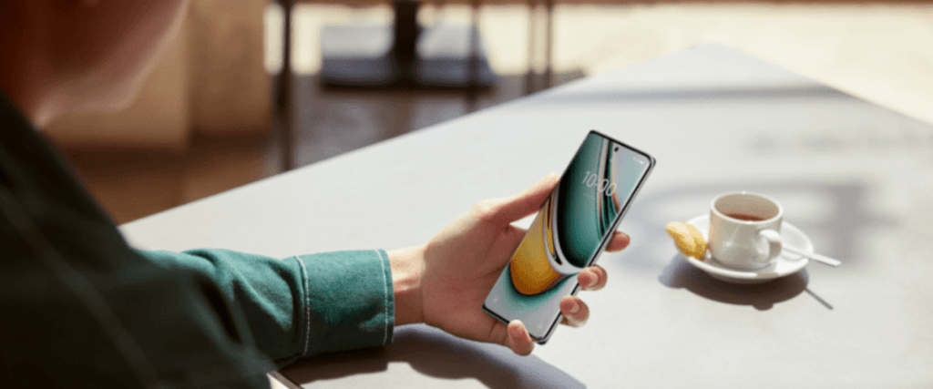 grafika przestawiająca nowoczesnego smartfona trzymanego w lewej dłoni na tle stołu