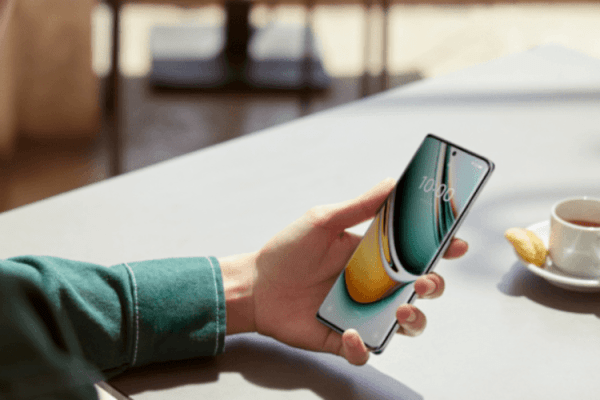 grafika przestawiająca nowoczesnego smartfona trzymanego w lewej dłoni na tle stołu