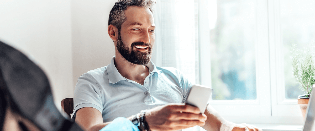 grafika przedstawiająca uśmiechniętego mężczyznę z brodą trzymającego smartfona i patrzącego się ekran monitora