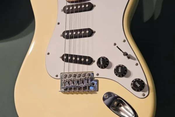 zdjęcie przedstawiające wiszącą na zielonej ścianie kremową gitarę elektryczną typu Fender Stratocaster z białą płytką maskującą